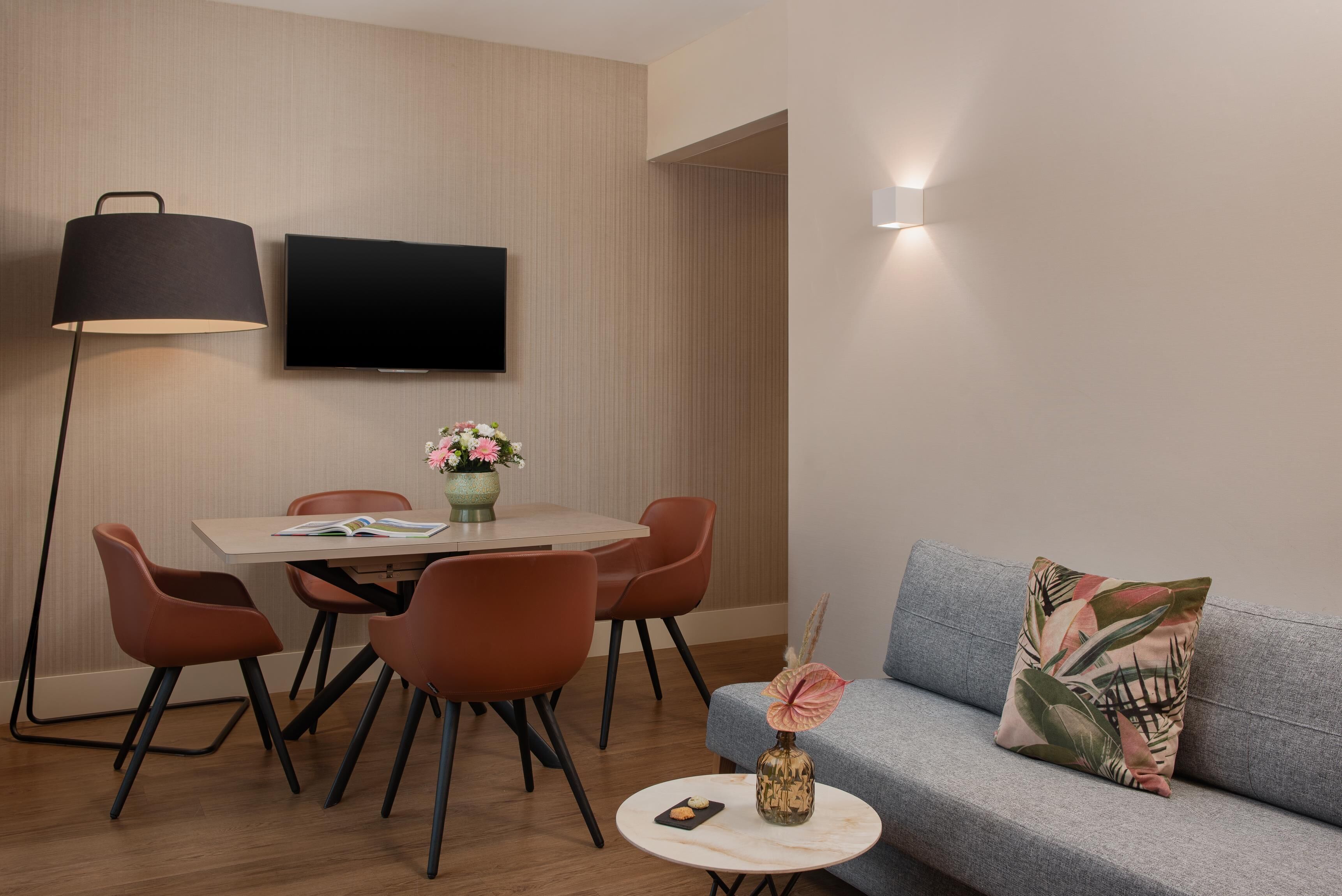 En moderne stue med et fladskærms-tv, et spisebord med stole, en sofa med en dekorativ pude, en gulvlampe og et lille bord med en vase og en bog.