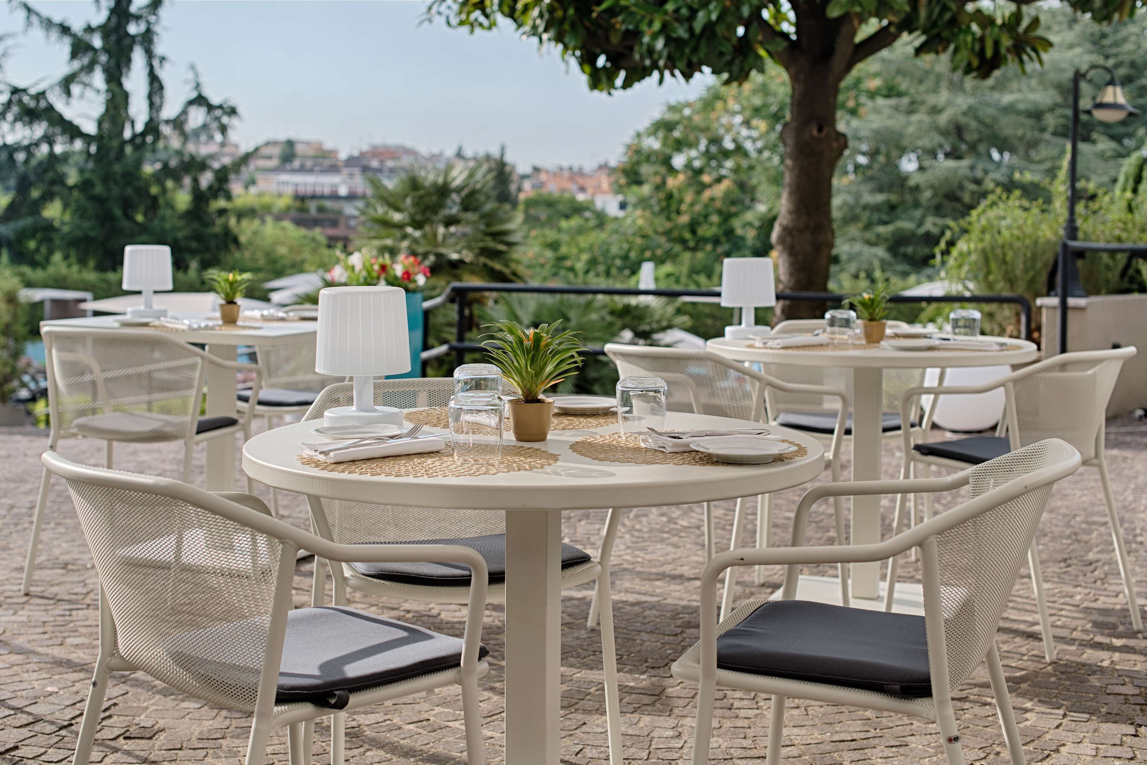 An outdoor patio with round tables and white wicker chairs, each with a small potted plant and a white table lamp.