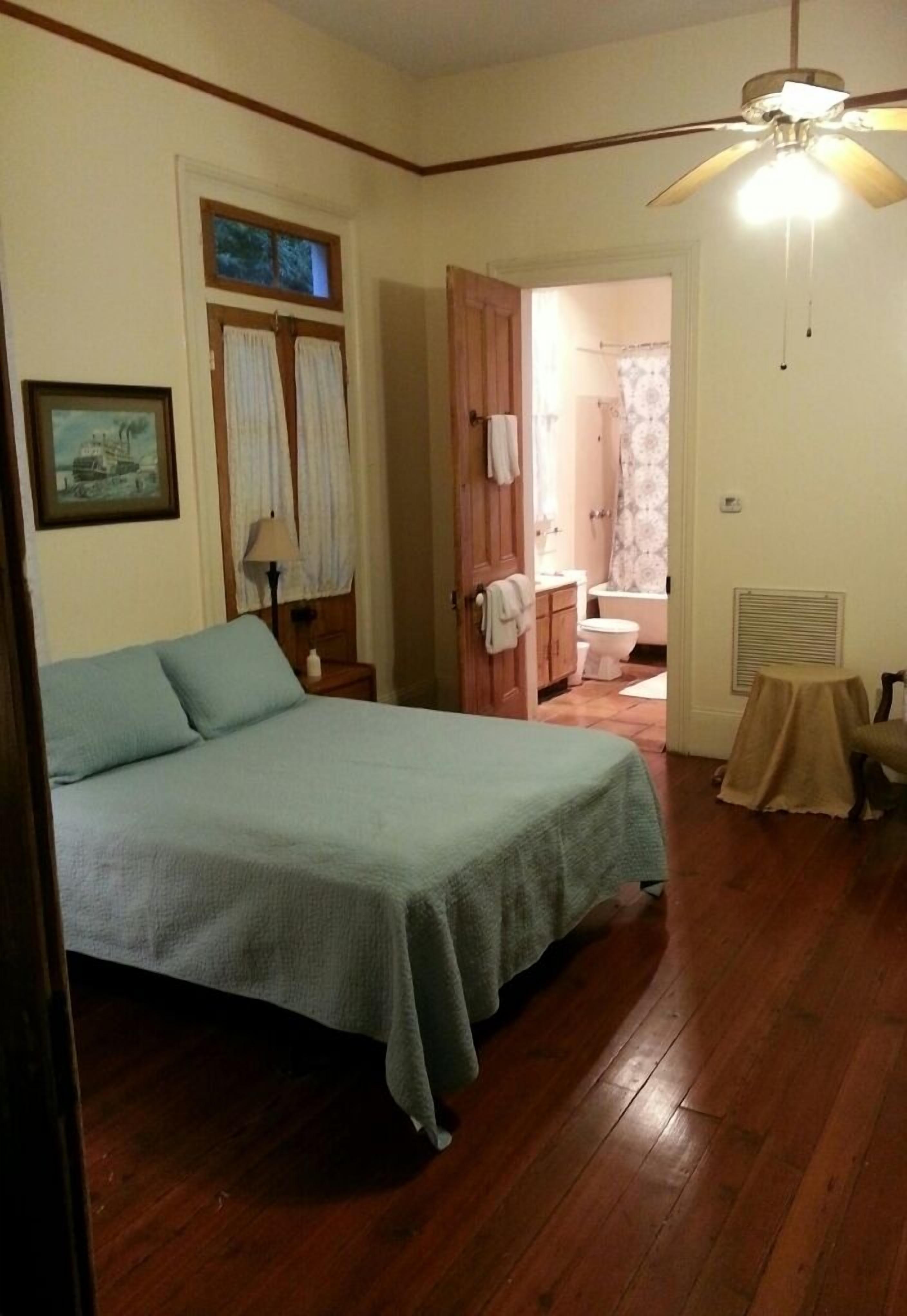 A bedroom with a bed, wooden flooring, a ceiling fan, a window with curtains, and a bathroom visible through an open door.