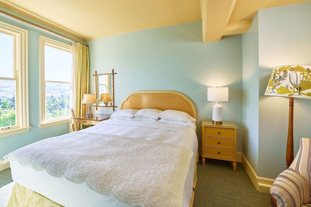 A bedroom with a bed, bedside tables, a lamp, a chair, and a view of the outdoors.