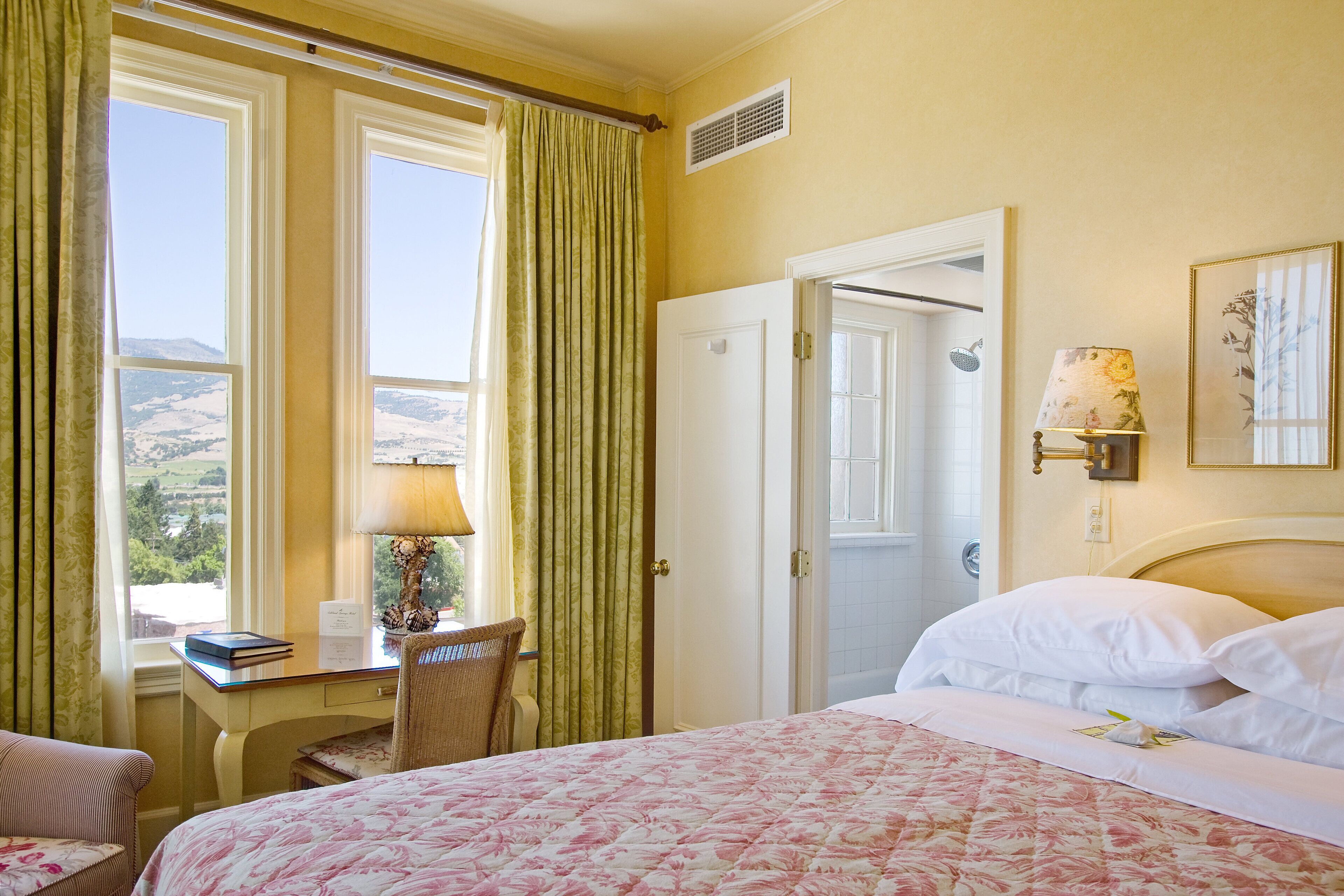 A hotel room with a bed, a desk, a chair, a window with curtains, and a door.