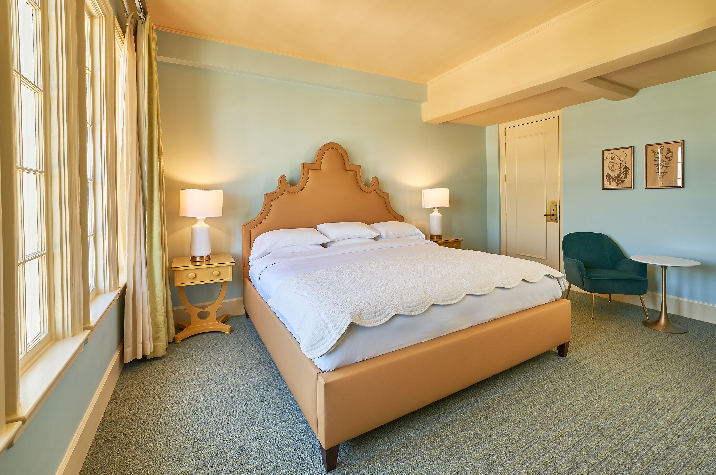 A neatly made bed with a wooden headboard, two bedside lamps, a small bedside table, a green armchair, and a round table.