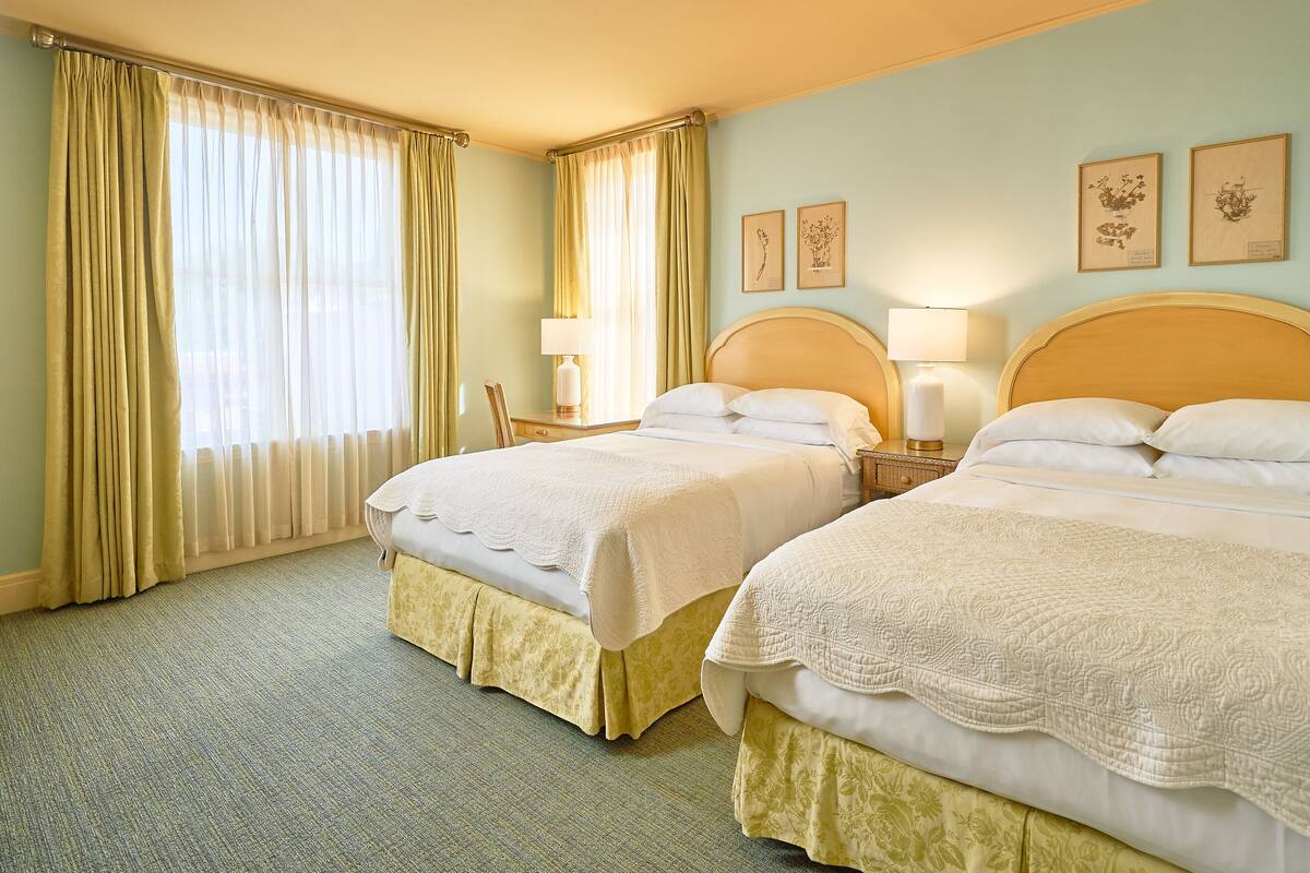 Habitación de hotel con dos camas, una ventana con cortinas, una mesita de noche con lámpara y cuadros en la pared.