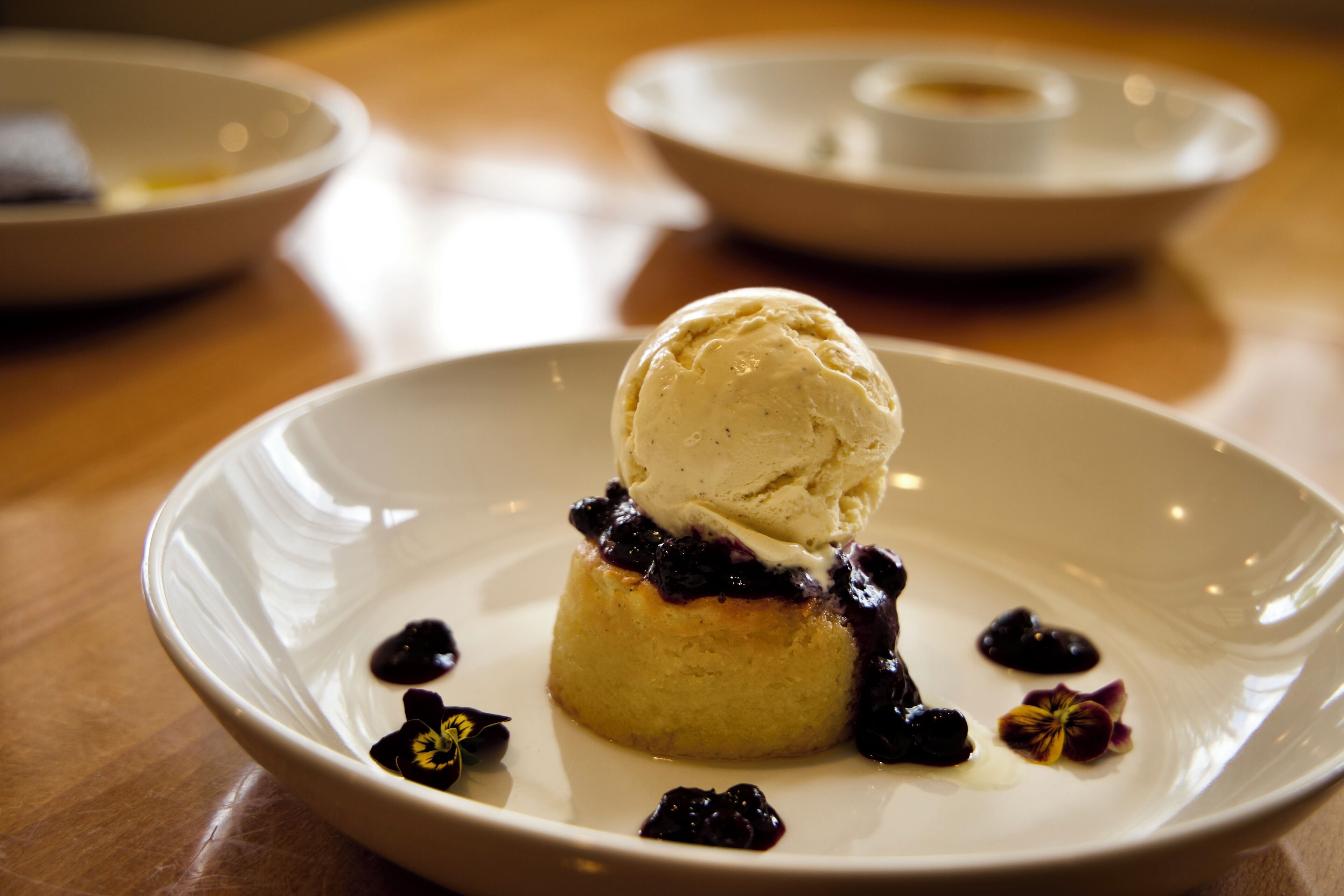 Postre sobre un plato blanco con una bola de helado, rodeado de bayas y una pequeña flor.