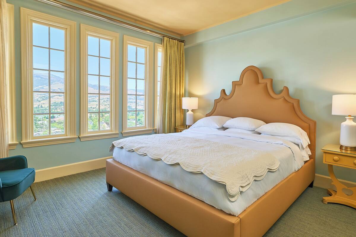 Un dormitorio con una cama grande, dos lámparas de noche, una silla y vistas al exterior a través de tres ventanas.