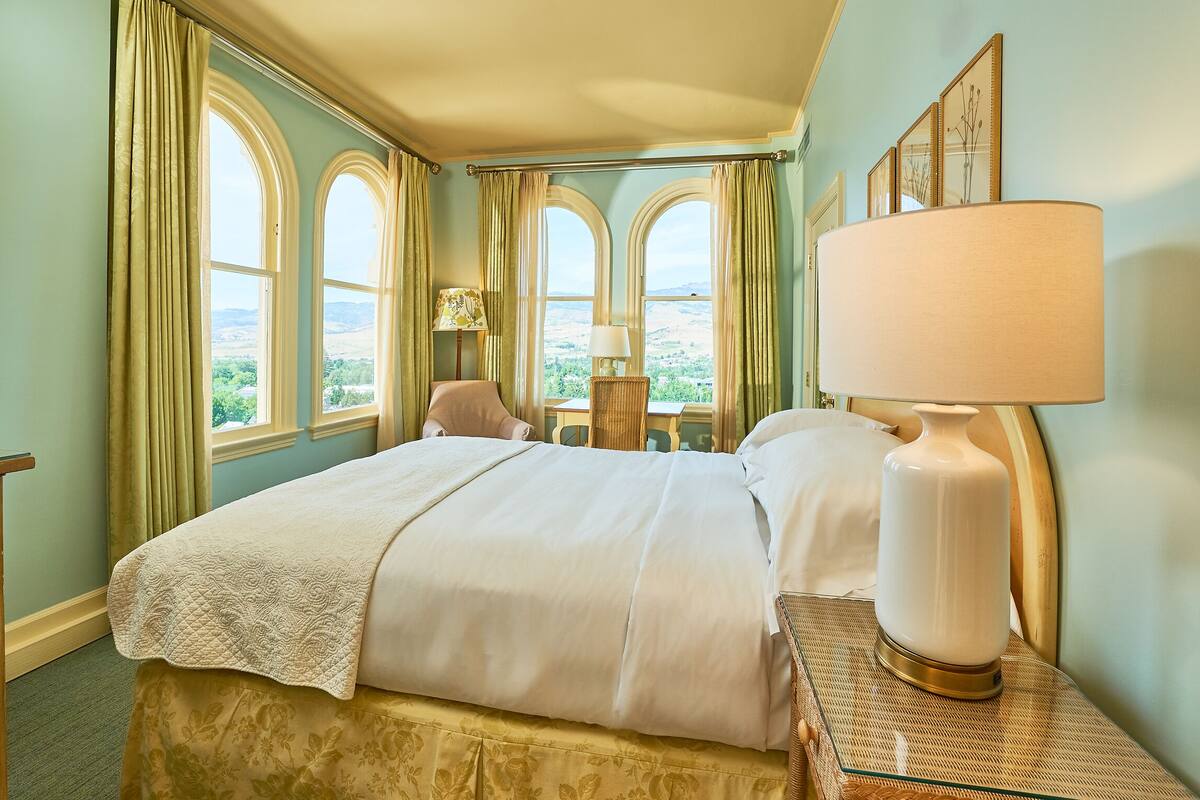 Habitación de hotel con una cama grande, una mesita de noche con lámpara, una silla y vistas del paisaje a través de ventanales arqueados.