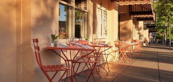 Des tables et des chaises sont disposées à l’extérieur, le long d’un trottoir, avec un bâtiment en arrière-plan.