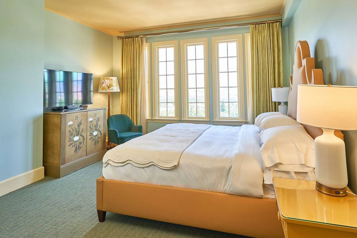 Habitación de hotel con cama, televisión, escritorio, silla y ventana con cortinas.