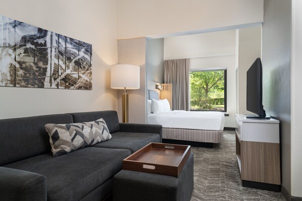 A hotel room with a sofa, a bed, a TV, a bedside table, and a window with a view of greenery. - SpringHill Suites by Marriott West Mifflin (West Mifflin)