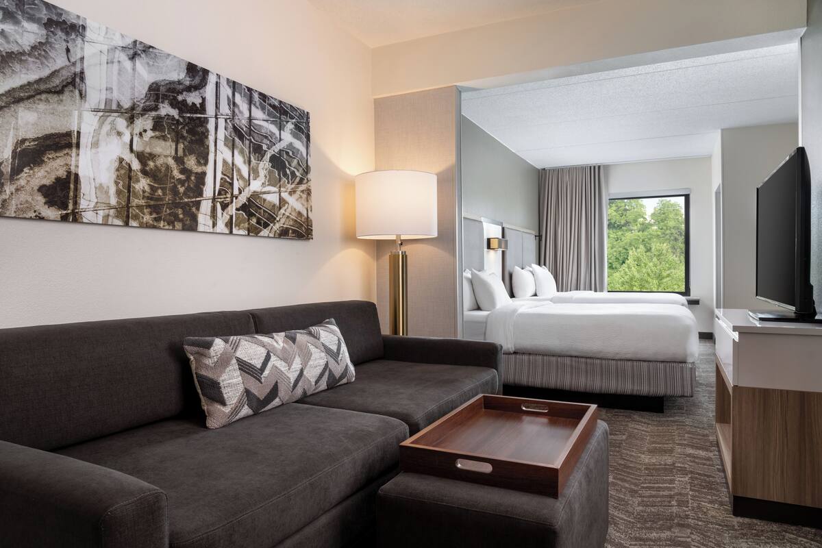 A hotel room with a sofa, a bed, a bedside table, a lamp, a TV, and a large abstract painting on the wall.