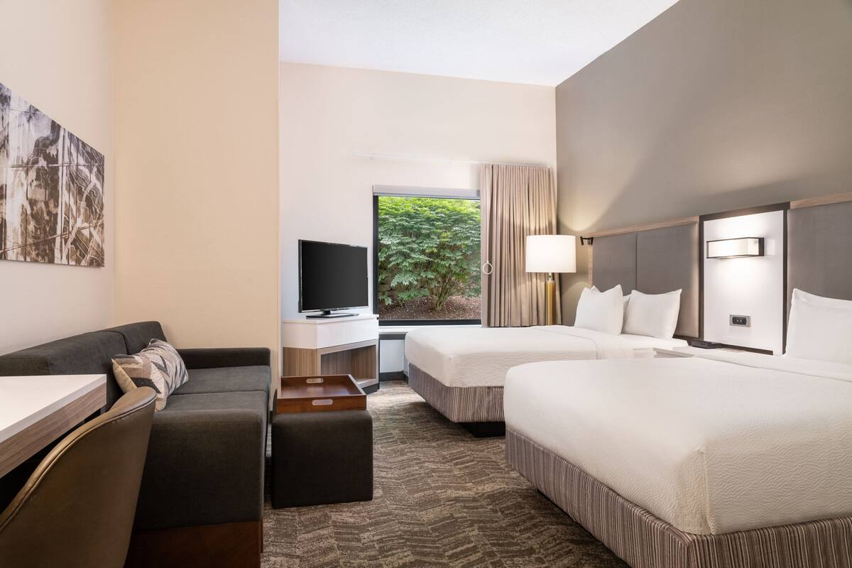 A hotel room with a sofa, a TV, two beds, a desk, and a window with a view of greenery.
