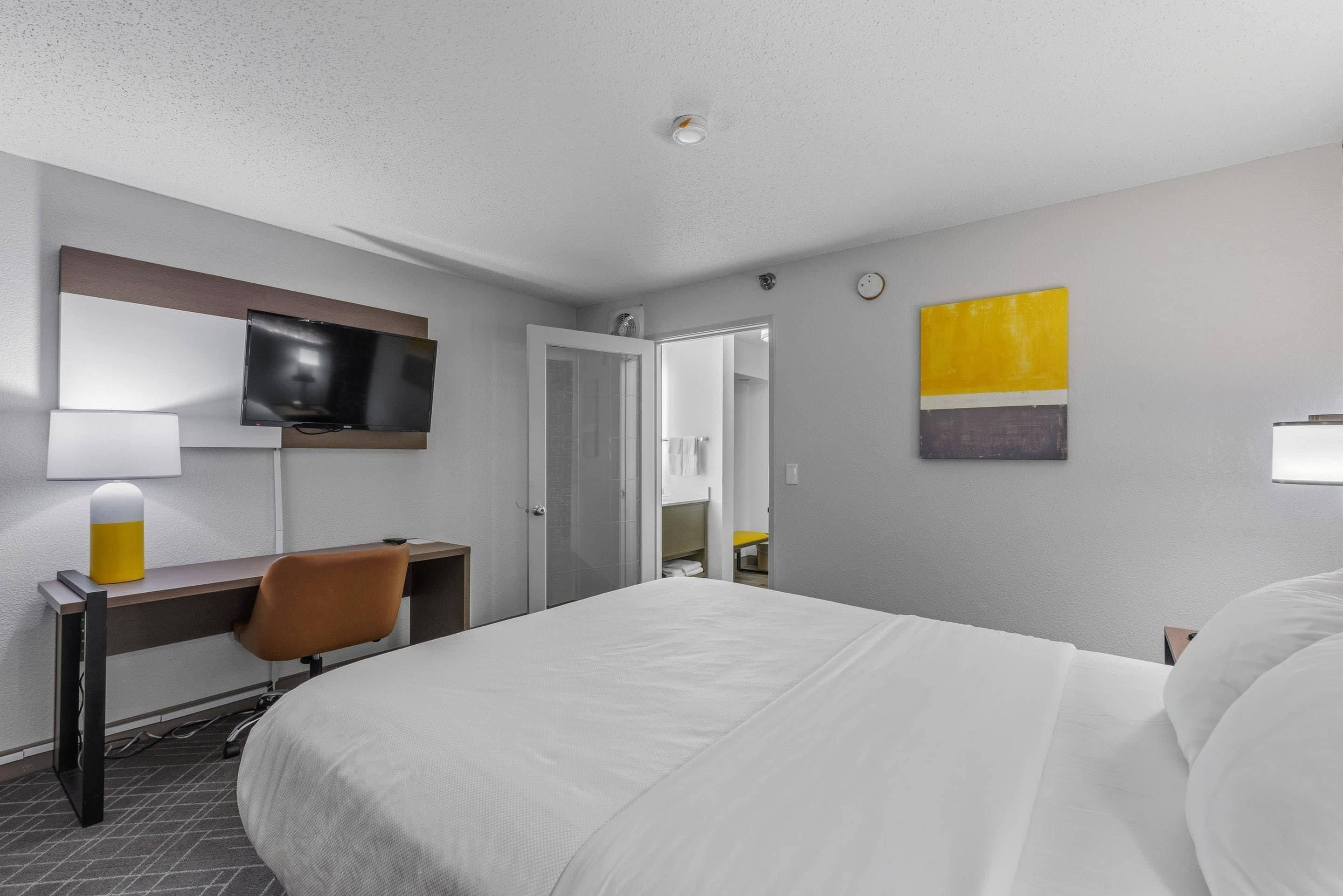 Camera d'albergo con un letto, una scrivania con una sedia, una televisione e un bagno visibile tramite una porta in vetro.
