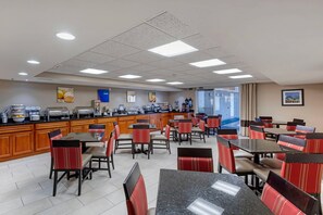 A hotel dining area with multiple tables and chairs, a kitchen counter with food service equipment, and a view of the outside through large windows. - Comfort Inn & Suites Jackson - West Bend (Jackson)