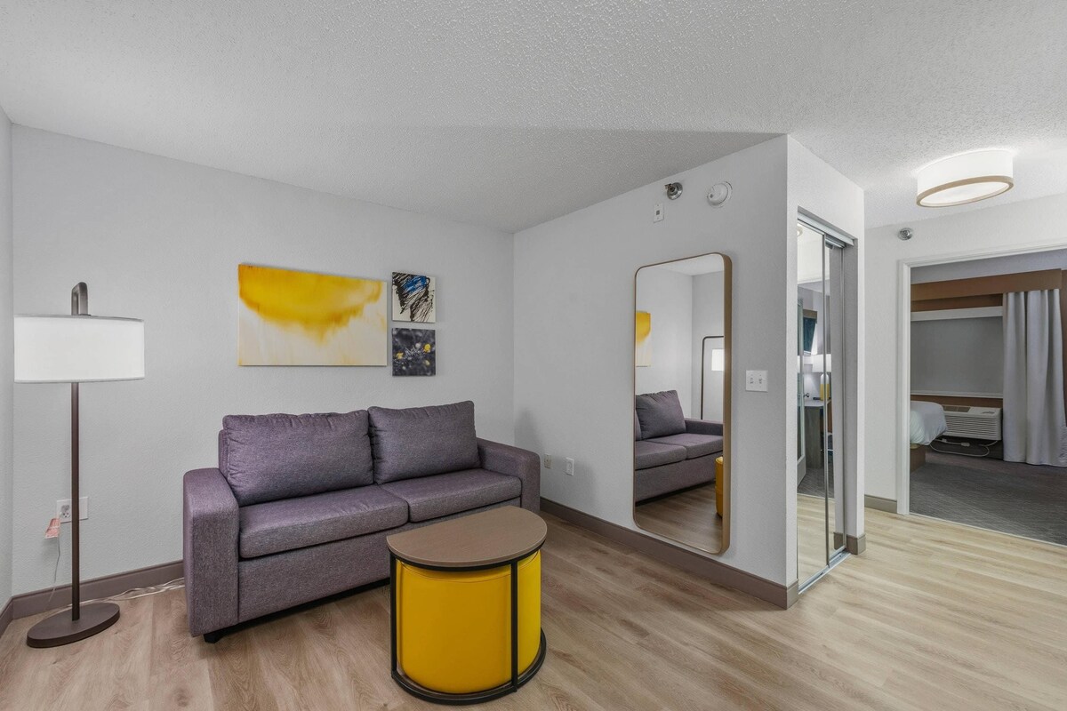 A modern hotel room with a sofa, a small round table, a floor lamp, and artwork on the wall.