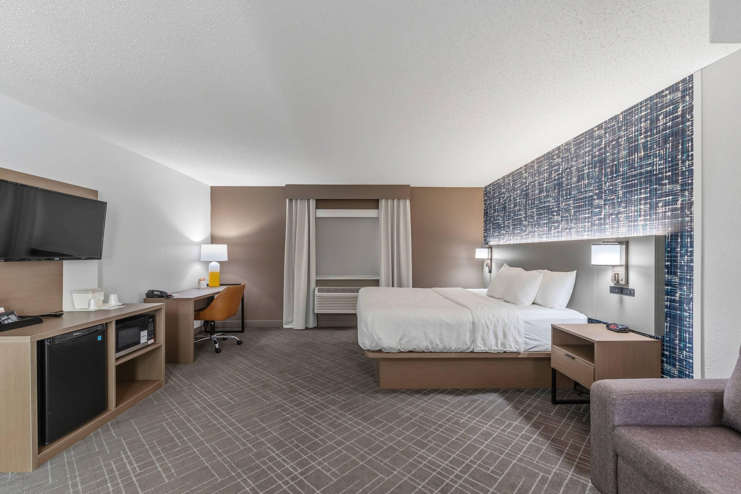 A hotel room with a bed, desk, chair, television, and a large abstract wall art piece.