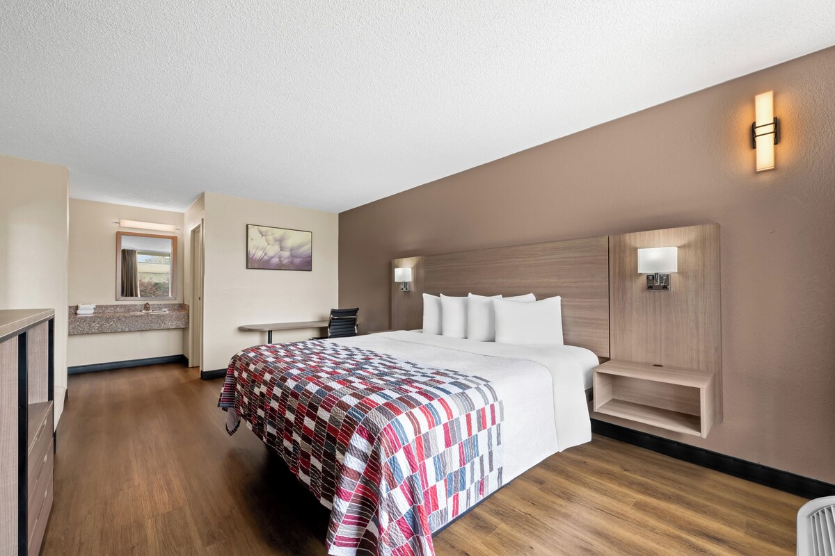 A hotel room with a large bed, a bedside table, a chair, a small desk, a mirror, and a wall-mounted shelf.