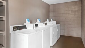Une buanderie équipée de quatre machines à laver et de quatre sèche-linge blancs, d’un panneau de commande et d’un mur en briques.