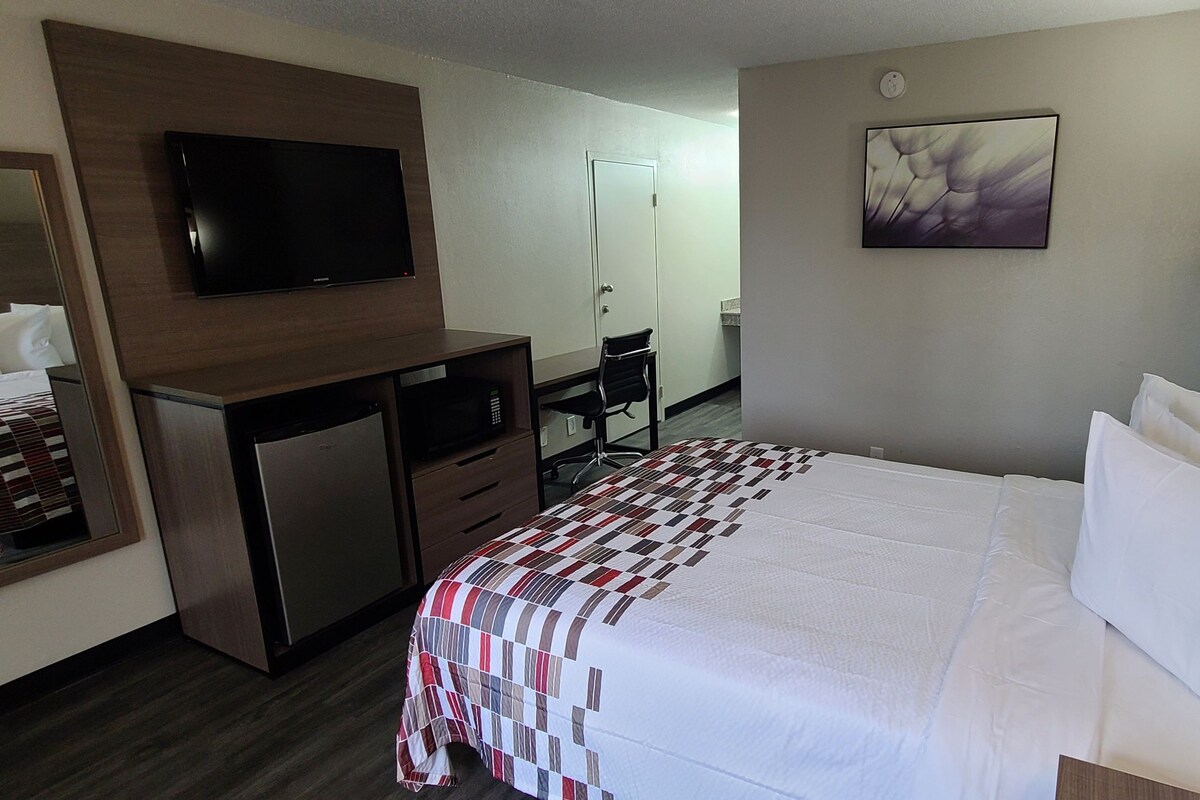 A hotel room with a bed, a desk, a chair, a television, and a painting on the wall.