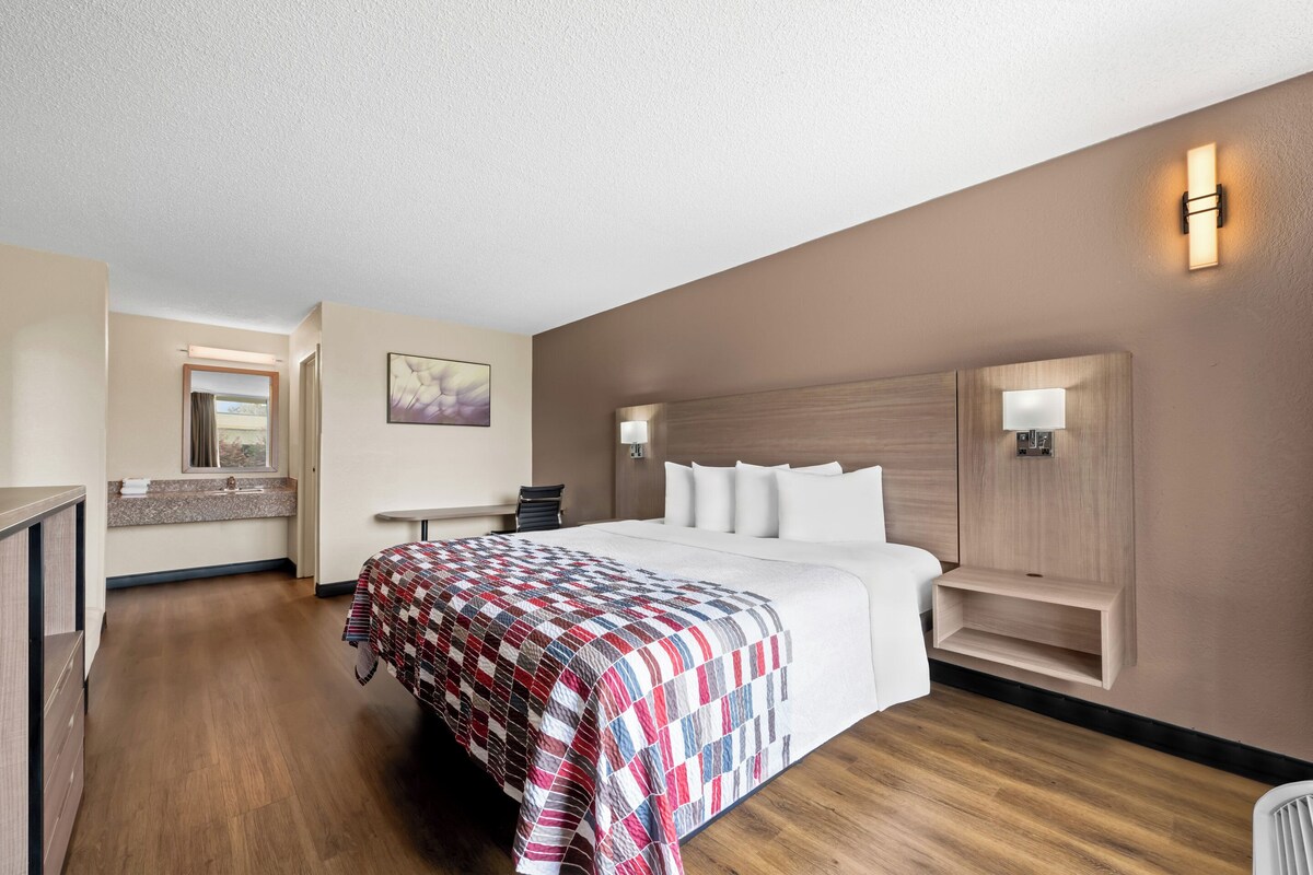 Une chambre d’hôtel avec un grand lit, une table de chevet, une chaise, un petit bureau, un miroir et une étagère fixée au mur.