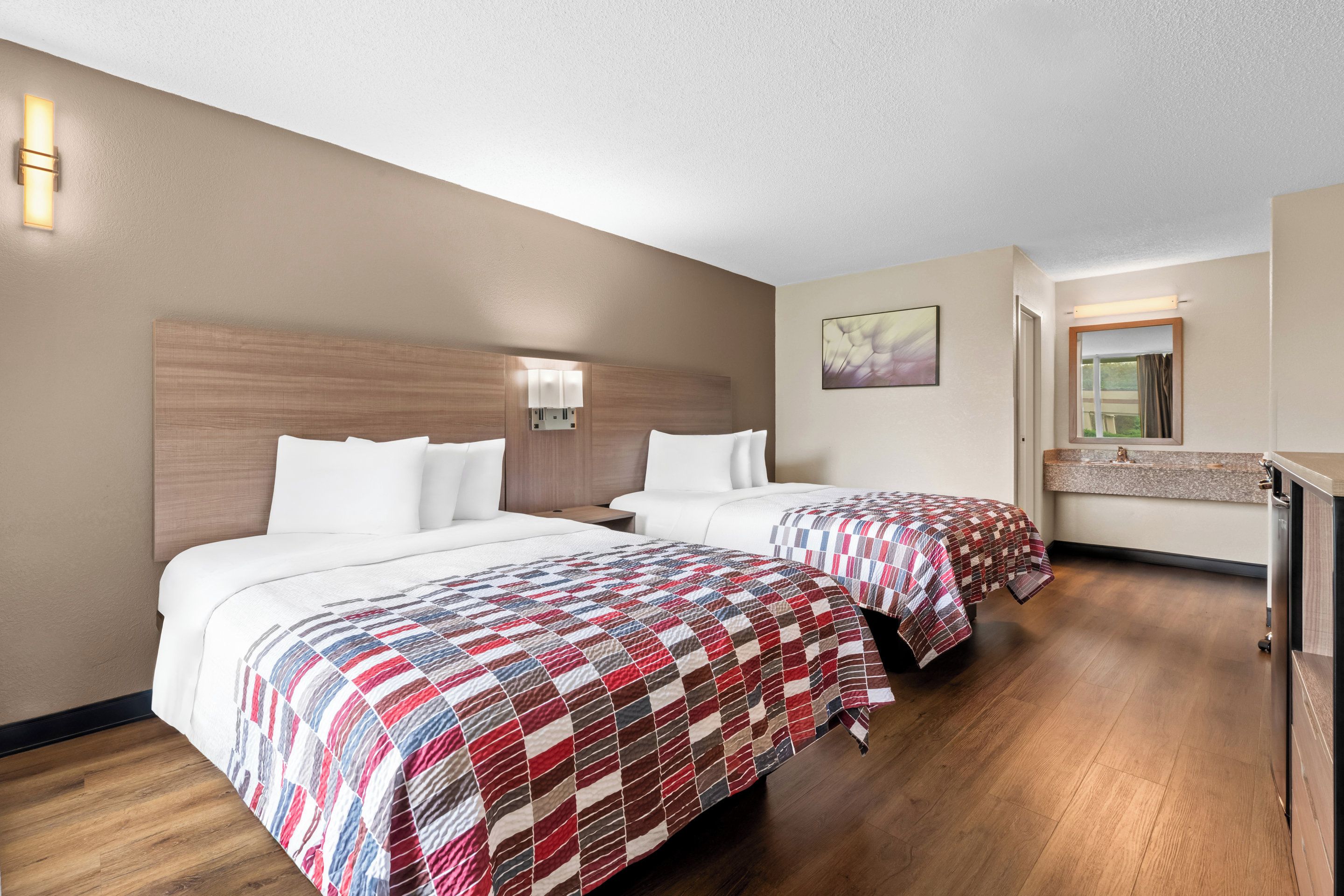 A hotel room with two beds, a wooden headboard, a nightstand, a bathroom with a mirror, and a painting on the wall.