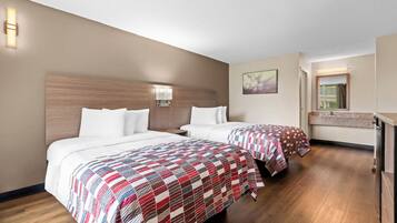 A hotel room with two beds, a wooden headboard, a nightstand, a bathroom with a mirror, and a painting on the wall.