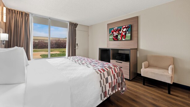 A hotel room with a large bed, a TV mounted on the wall, a chair, and a view of the outdoors. - Red Roof Inn Charlotte - Airport (Charlotte)