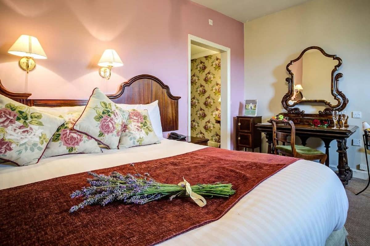 A neatly made bed with floral pillows, a red throw blanket, and a bouquet of lavender.