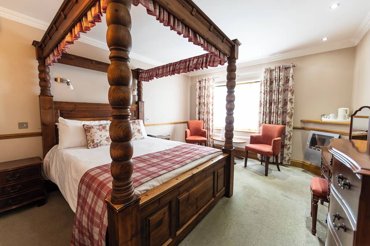 A four-poster bed with a wooden frame, a red and white checkered bedspread, and a floral pillow. There is a wooden nightstand with a lamp, a wooden dresser, and a chair with a floral curtain.
