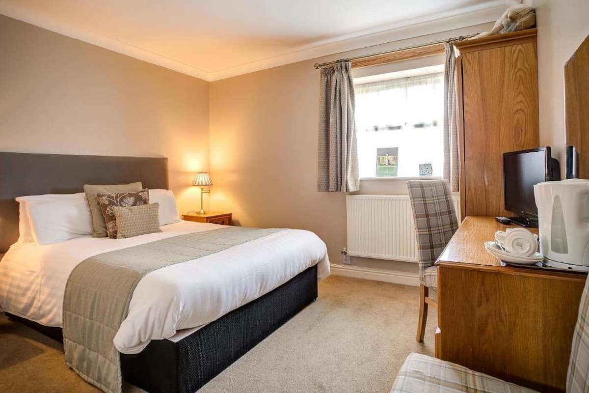 A hotel room with a bed, bedside table, lamp, window with curtains, radiator, and a desk with a computer and coffee maker.