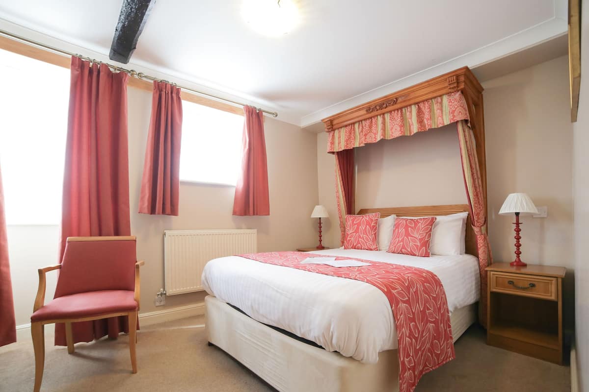 A bedroom with a four-poster bed, a chair, a bedside table with a lamp, and a window with curtains.