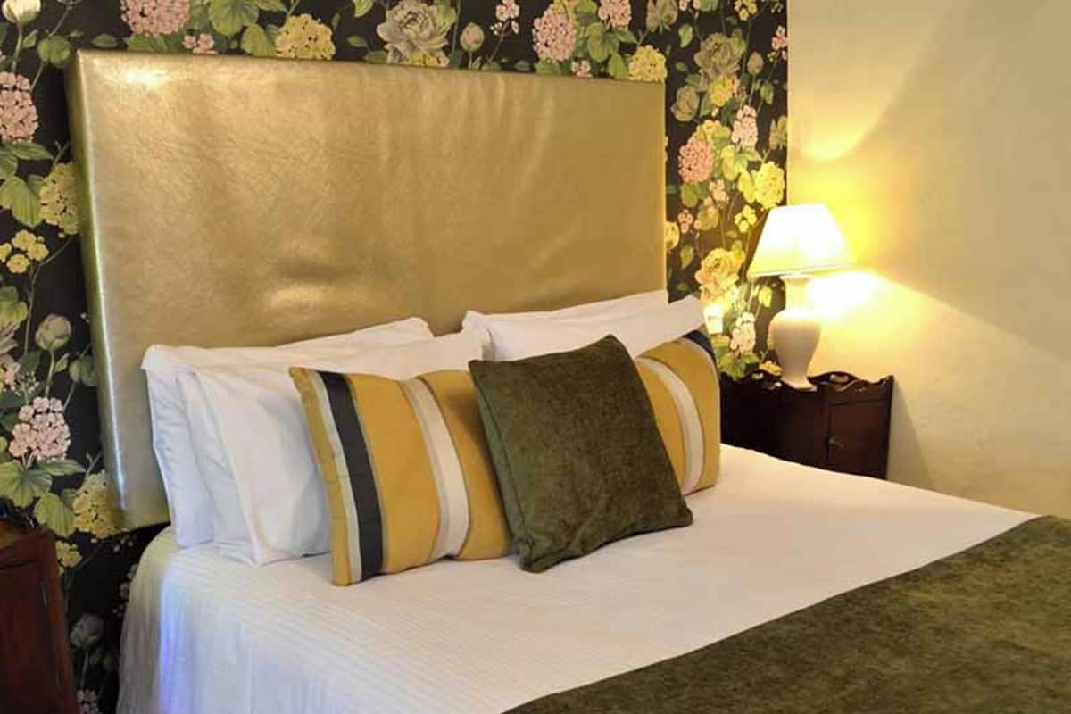 A hotel room with a large bed, multiple pillows, a nightstand with a lamp, and floral wallpaper.
