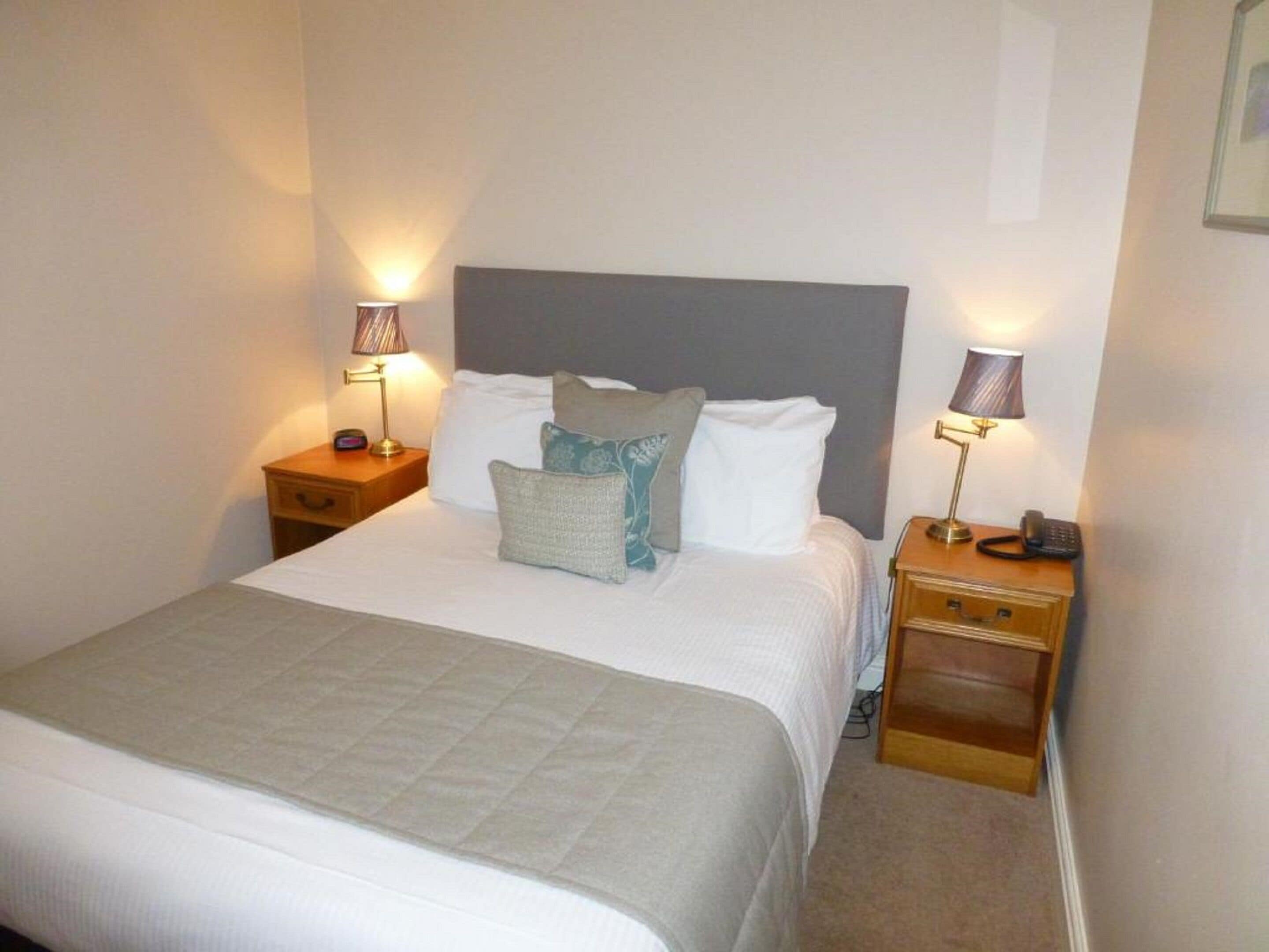 A neatly made bed with a grey headboard, two bedside tables with lamps, and a small wooden nightstand with a phone.