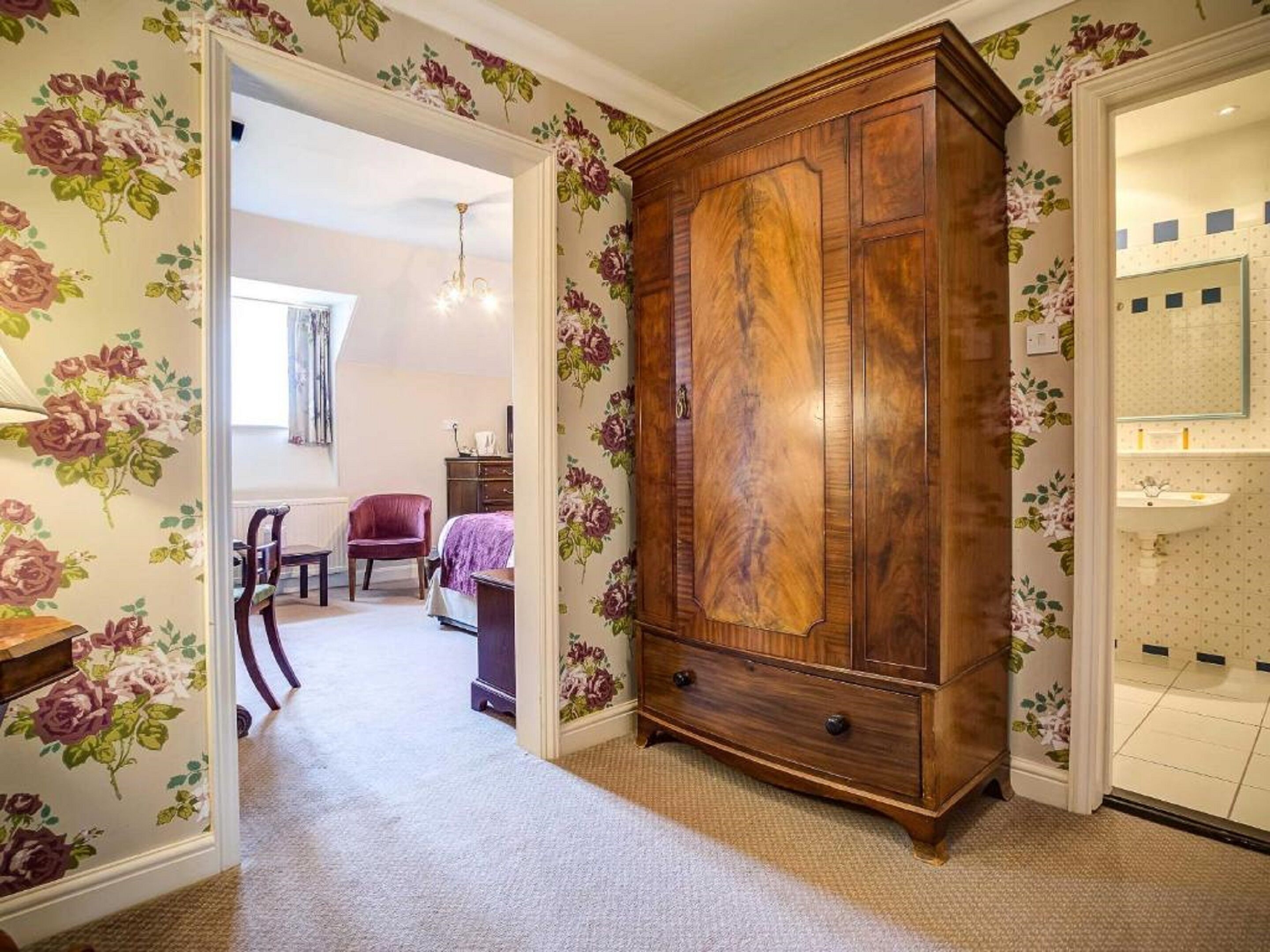 Un dormitorio con papel tapiz floral, un armario de madera, una cama y un baño con espejo y lavamanos.