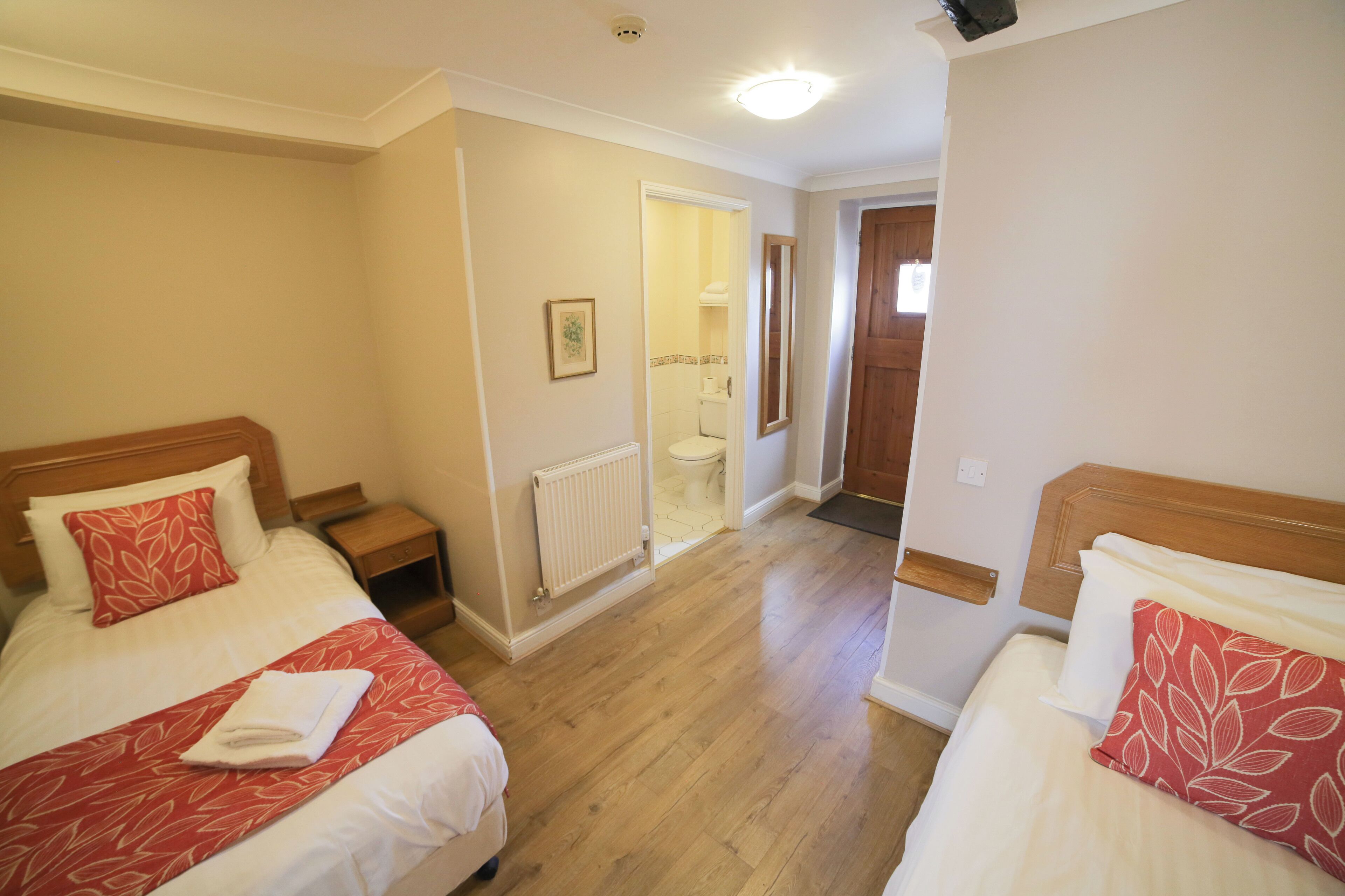 A hotel room with two beds, wooden headboards, a radiator, a wooden floor, and a door leading to a bathroom.