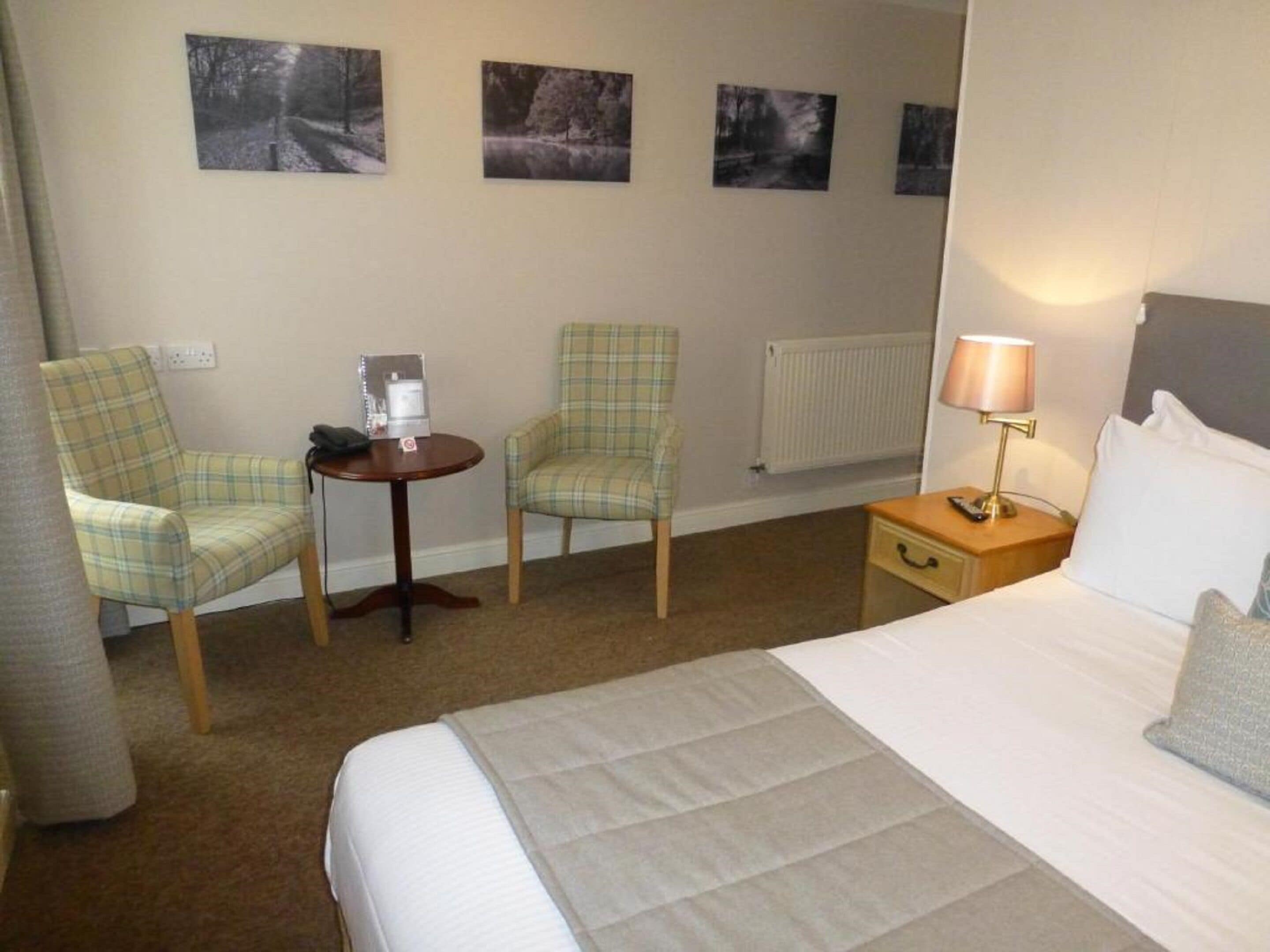 A hotel room with a bed, two chairs, a small table with a lamp, and three framed pictures on the wall.