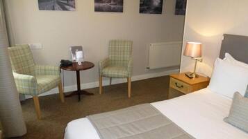 A hotel room with a bed, two chairs, a small table with a lamp, and three framed pictures on the wall.