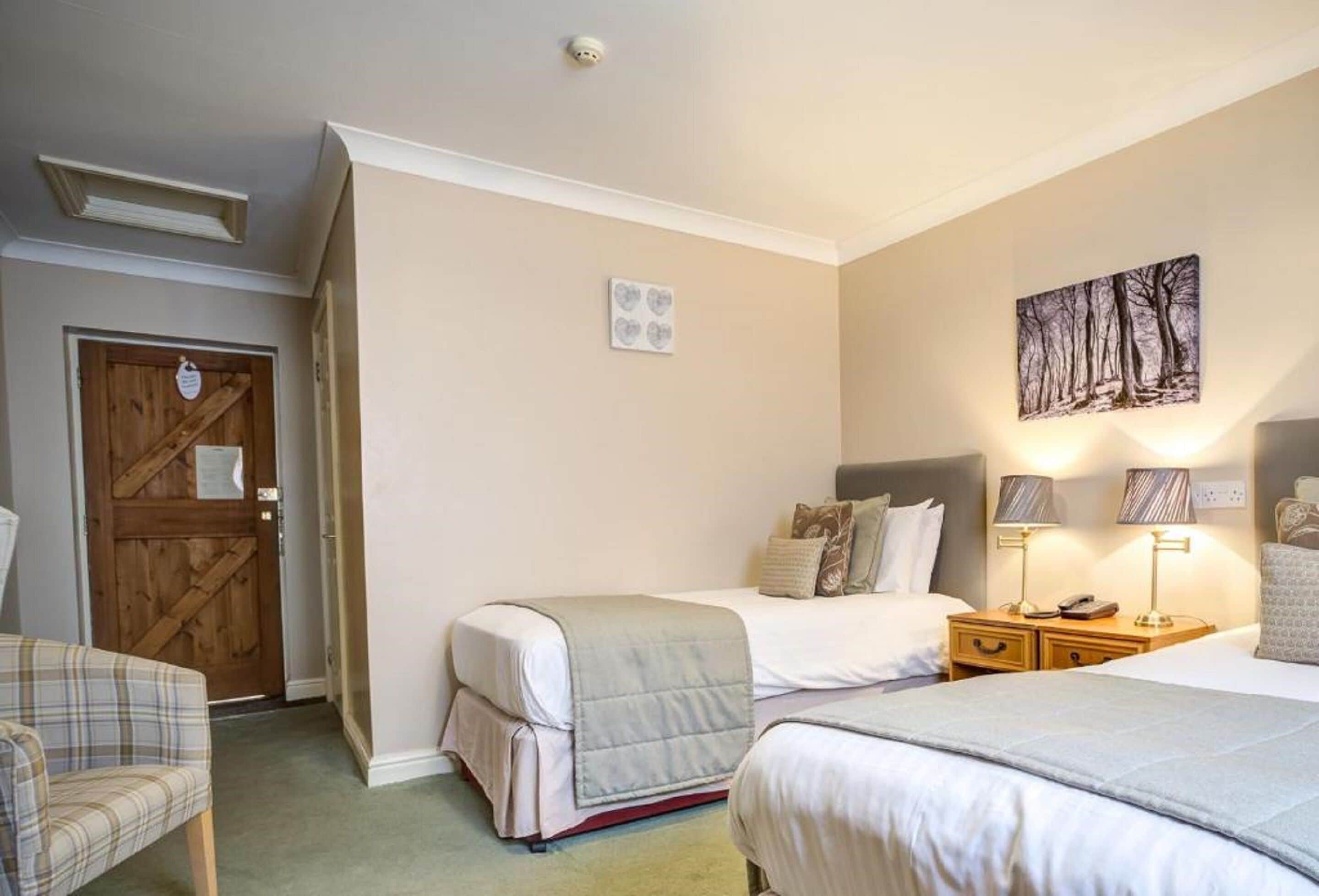 A bedroom with two beds, a wooden door, a chair, and a wall-mounted lamp.
