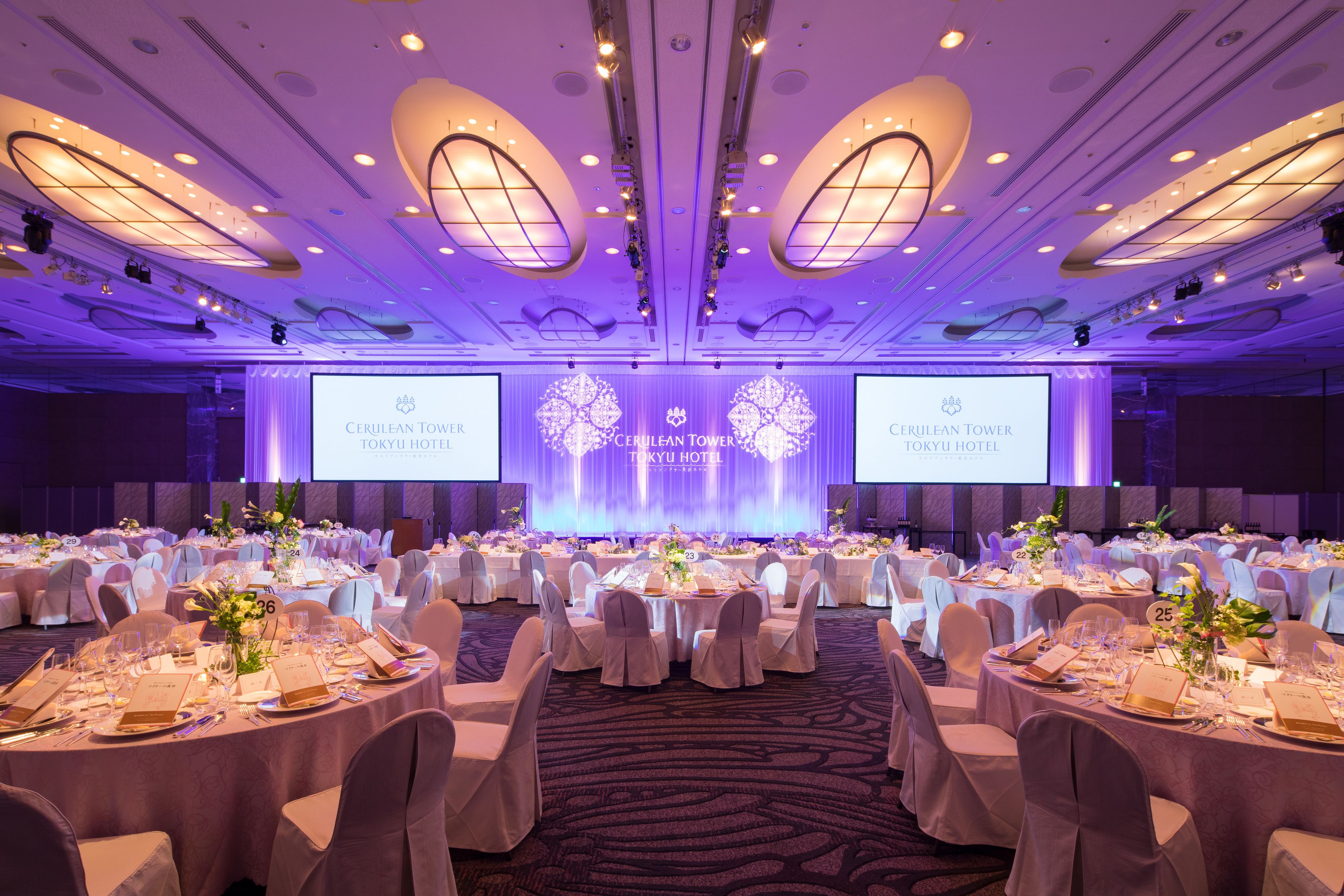 A large conference room with multiple round tables, each set with white tablecloths, chairs, and name cards.