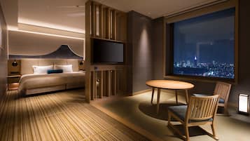 Quarto de hotel moderno com uma cama grande, televisão, área de refeições com cadeiras e vista para o horizonte noturno.