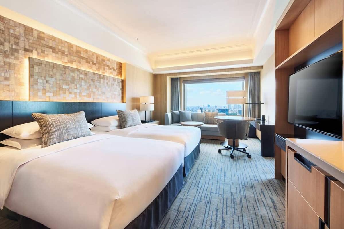 A hotel room with two beds, a desk, a TV, and a city view.