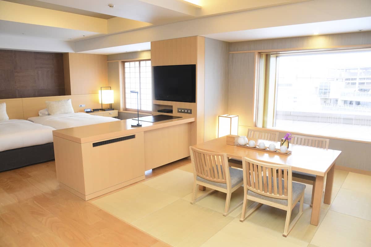 A hotel room with a bed, a desk with a TV, a dining table with chairs, and a large window with a city view.