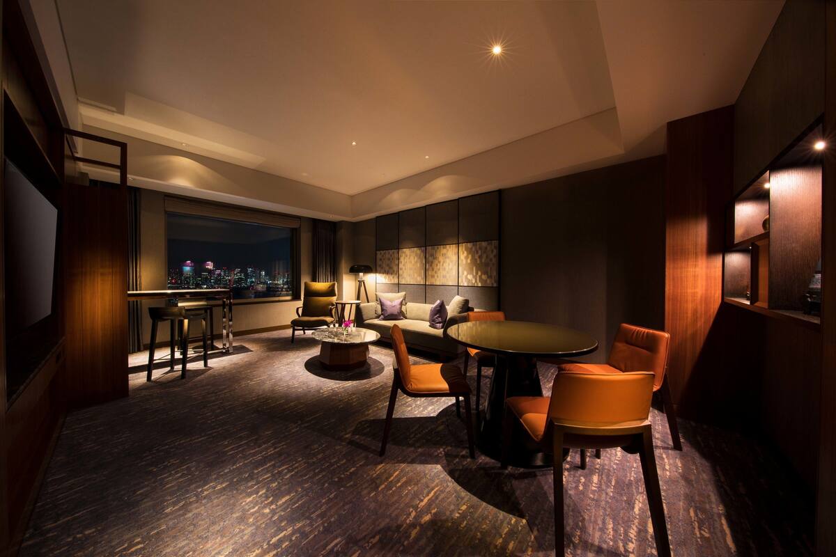 A modern hotel room with a large window offering a city view, a cozy seating area with a round table, and a bar area.
