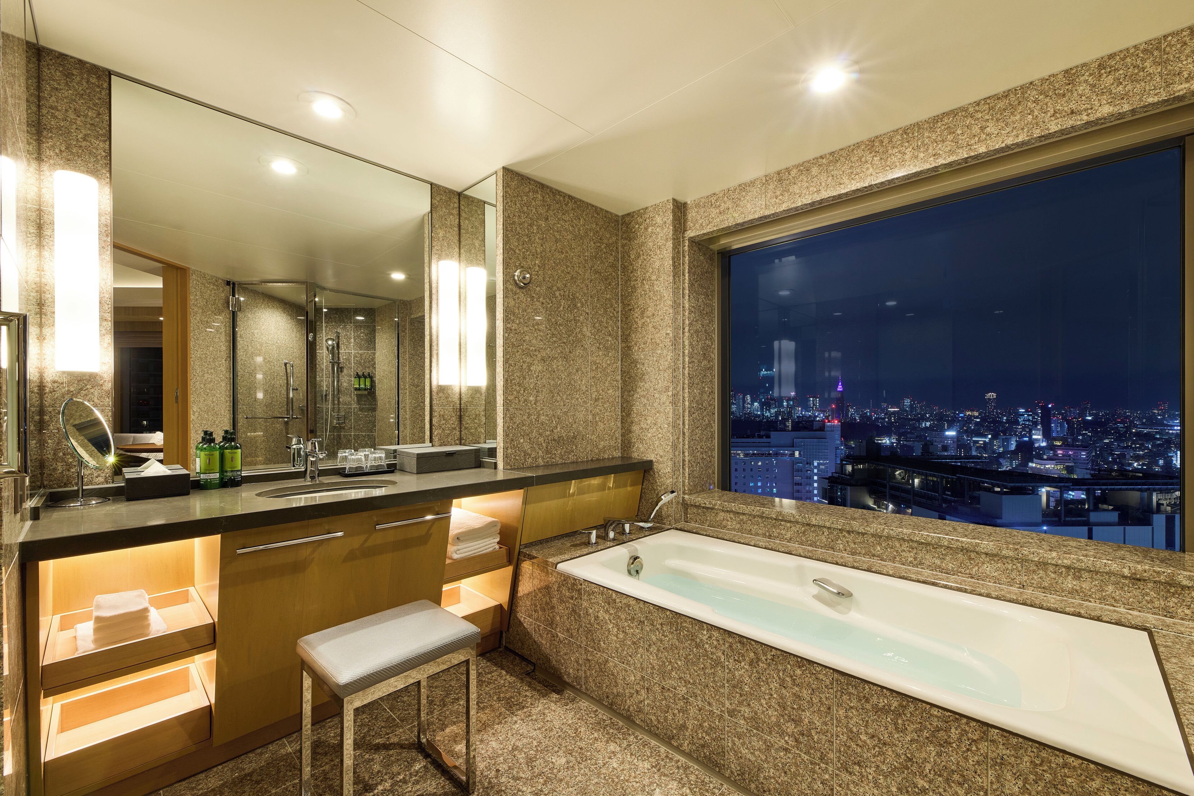Un baño moderno con una bañera grande, un tocador con espejo y vistas a la ciudad a través de un amplio ventanal.