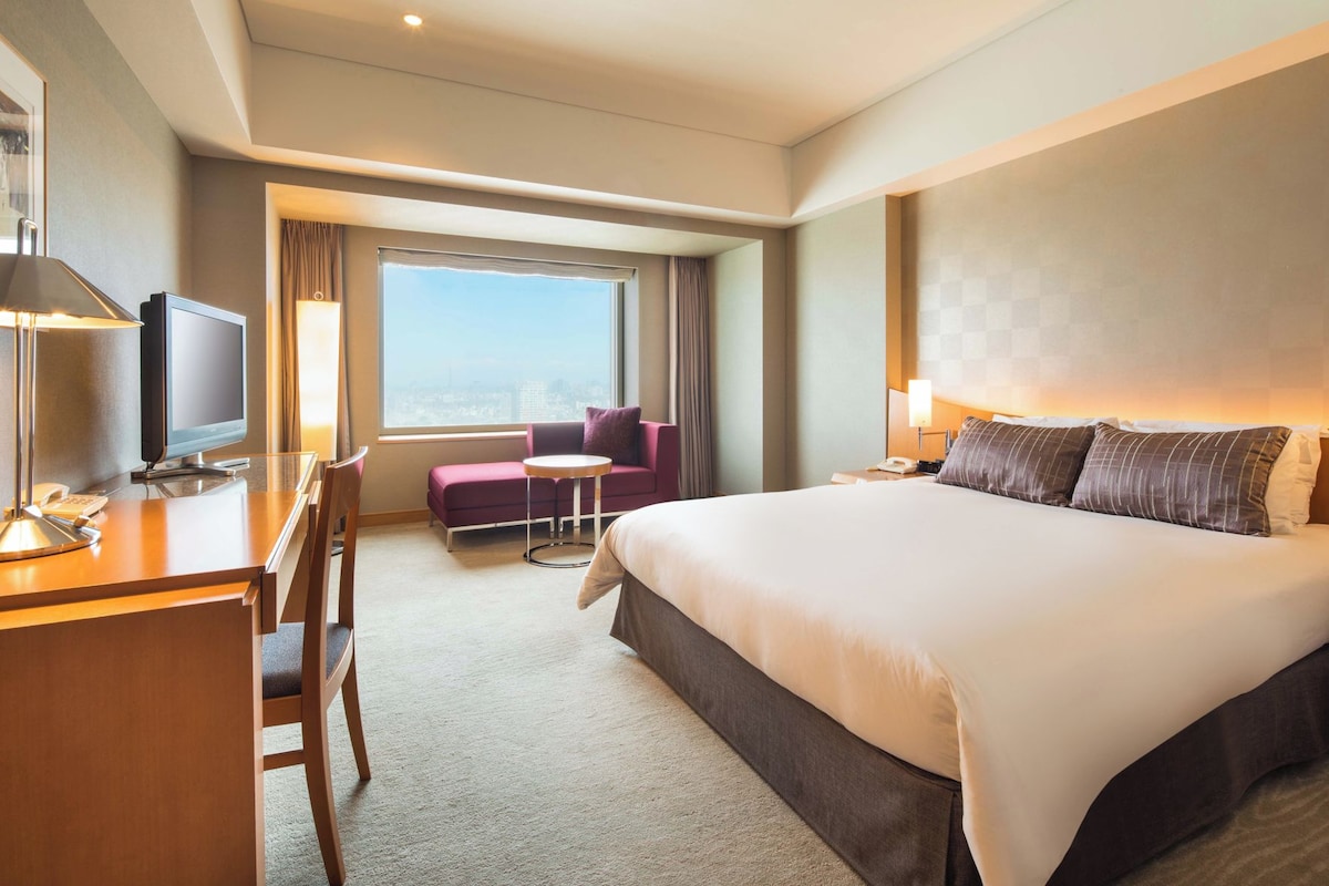 A hotel room with a large bed, a desk with a computer, a chair, a small table, and a window with a city view.