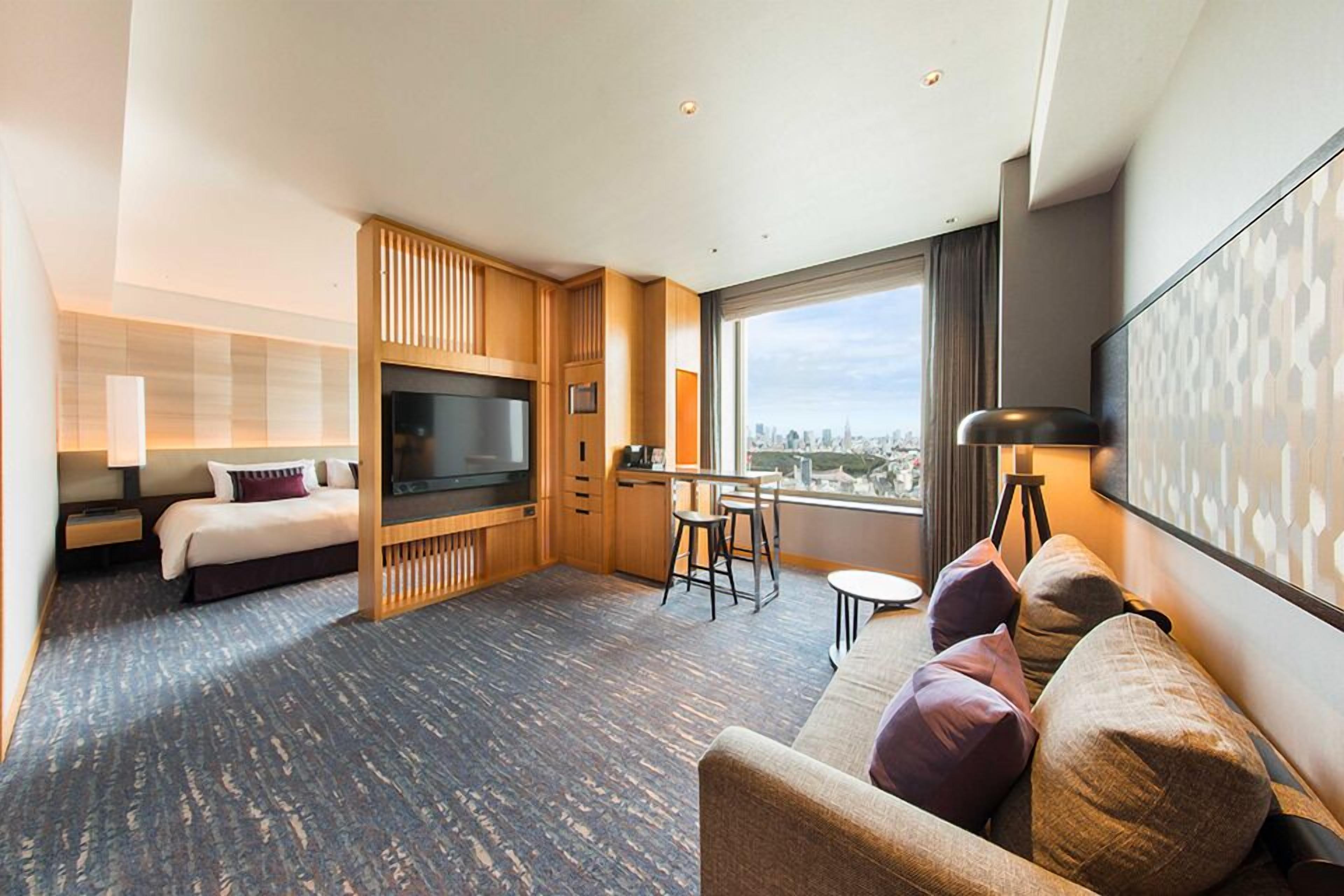 A hotel room with a bed, a TV, a dining area, and a large window with a city view.