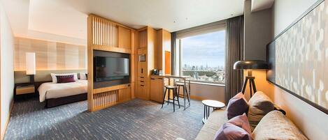 A hotel room with a bed, a TV, a dining area, and a large window with a city view.