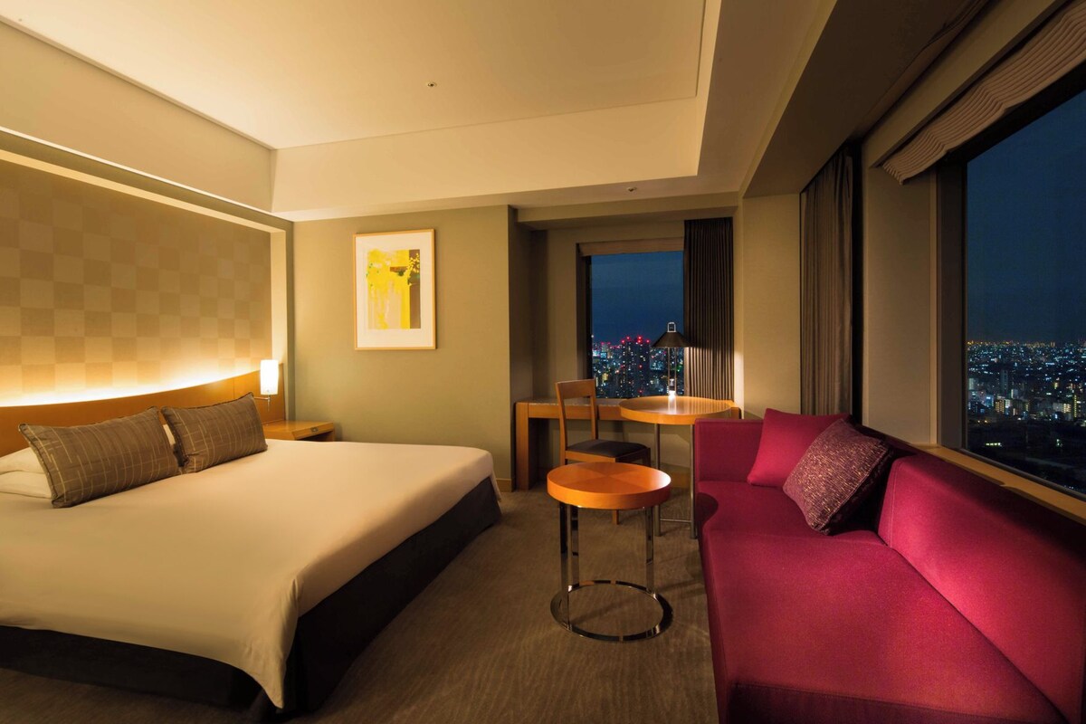 A hotel room with a bed, a red sofa, a small table, and a chair. There is a window with a city view.