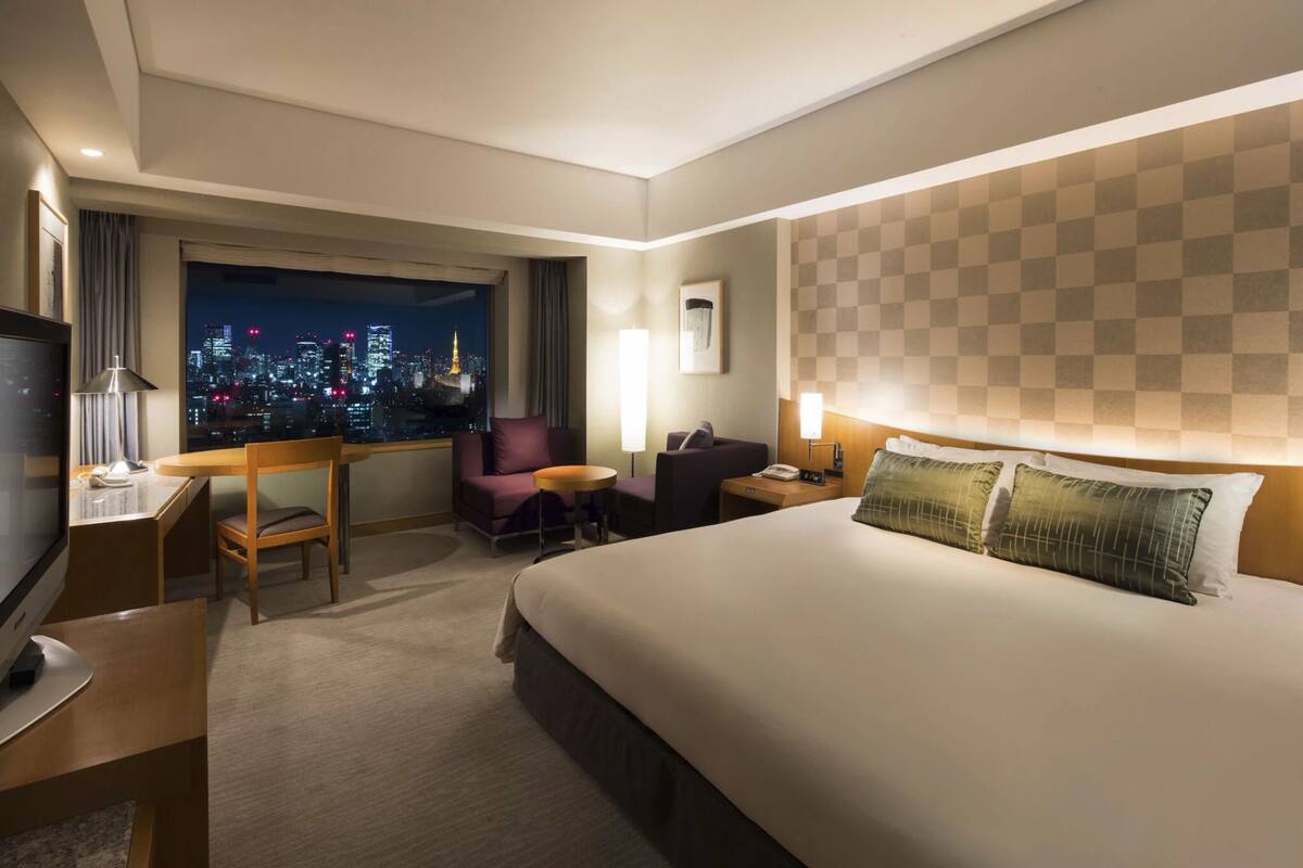 A hotel room with a large bed, a desk with a chair, a TV, and a view of a cityscape at night.