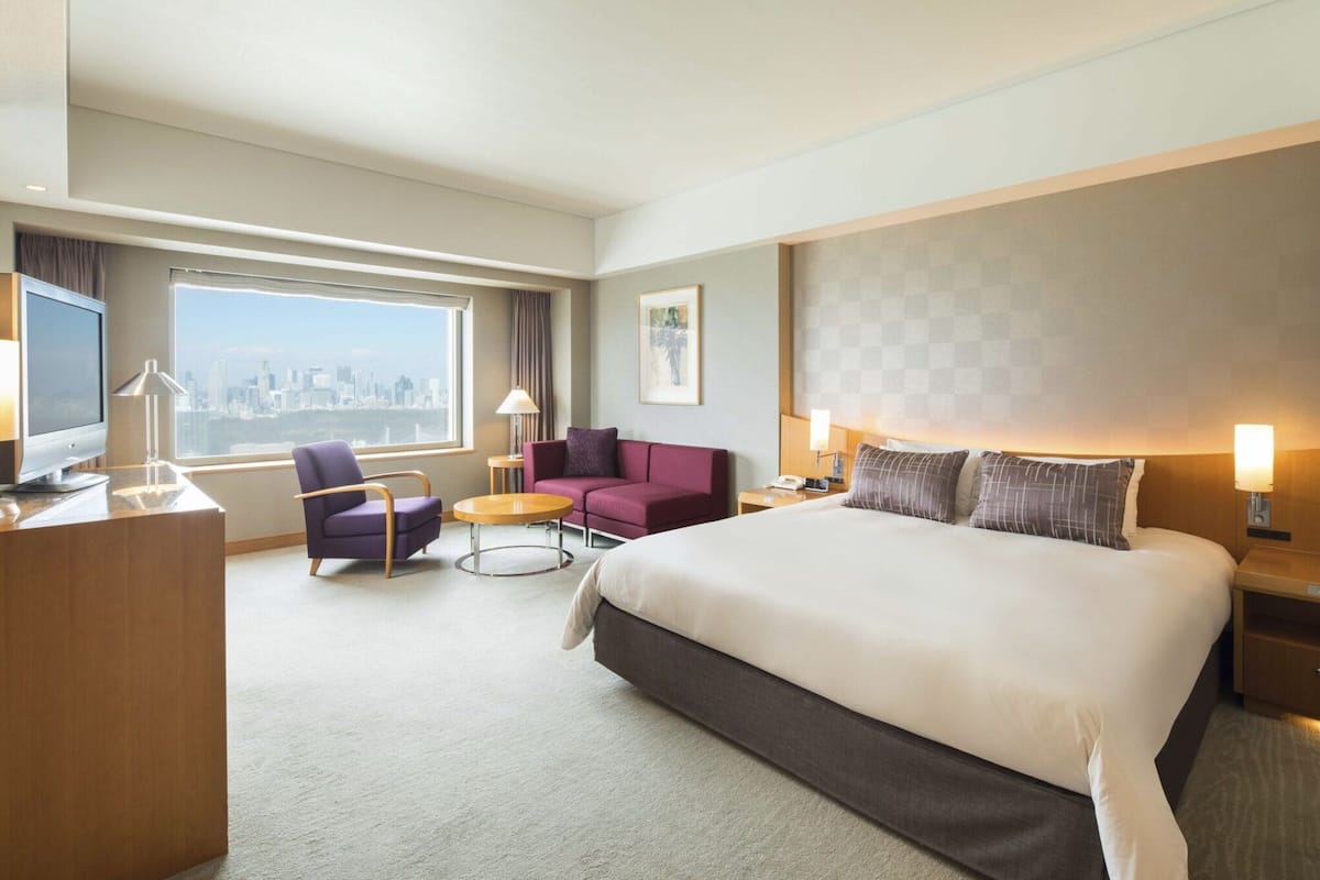 A hotel room with a large bed, a desk, a chair, a small table, and a view of a city skyline.