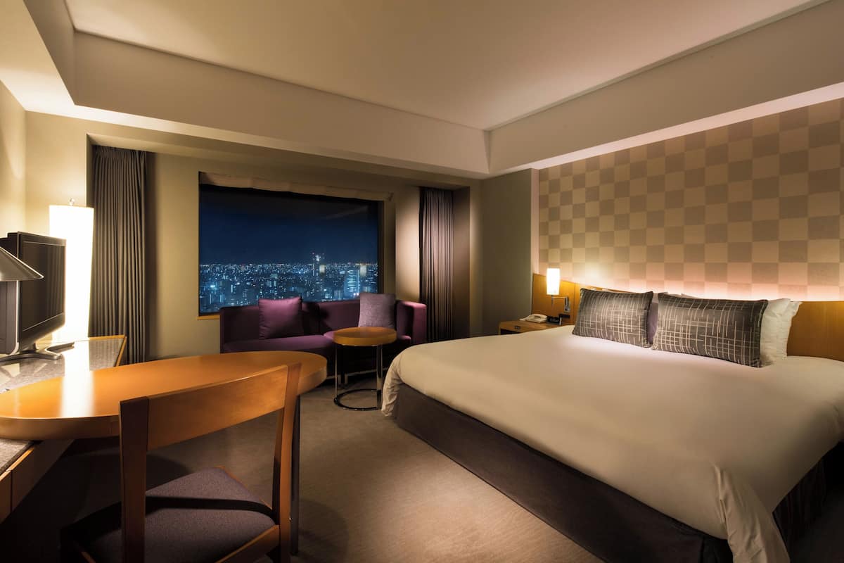 A hotel room with a large bed, a desk with a chair, a small round table, and a view of a cityscape at night.